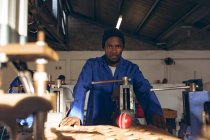 Front view close up of a young African American man leaning at a workbench in a cricket ball factory looking straight to camera. — Stock Photo