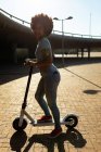 Vista lateral de perto de uma jovem mulher de raça mista montando uma scooter elétrica em um parque urbano, olhando para a câmera sorrindo, retroiluminado com brilho da lente — Fotografia de Stock