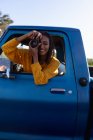 Gros plan d'une jeune femme métisse assise sur le siège passager avant d'une camionnette, penchée par la fenêtre latérale prenant des photos avec un appareil photo lors d'un voyage en voiture — Photo de stock