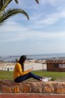 Vista laterale di una giovane donna di razza mista seduta fuori su un muro con uno smartphone, una palma e una vista sul mare sullo sfondo — Foto stock