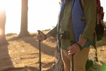 Side view mid section of man wearing a backpack, with a camera hanging on strap around his neck and holding Nordic walking sticks standing in the sun, during a hike — Stock Photo