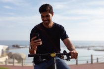 Вид спереду молодого змішаного чоловіка, який сидить на велосипеді, використовуючи смартфон у сонячний день, вид на море на задньому плані — стокове фото