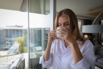 Vista frontale da vicino di una giovane donna caucasica che indossa una camicia bianca e beve caffè in un appartamento con gli occhi chiusi
. — Foto stock