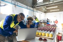 Front view close up of a young mixed race male manager standing and looking at a laptop with a young mixed race male worker sitting at a workbench with finished cricket balls at a cricket ball factory. — Stock Photo