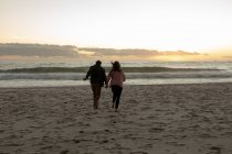 Задний вид зрелого кавказца мужчины и женщины, держащихся за руки и бегущих по пляжу к морю вместе на закате — стоковое фото