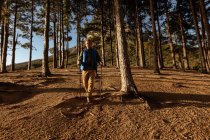 Vista frontal de un hombre caucásico maduro usando bastones nórdicos caminando a través de un bosque durante una caminata . - foto de stock