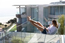 Vue latérale d'une jeune femme caucasienne portant une chemise blanche assise sur une chaise sur un balcon avec les jambes levées, tenant une tasse de café et regardant loin, mer, plage et bâtiments en arrière-plan . — Photo de stock