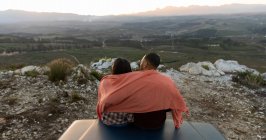 Vista posteriore di una giovane coppia mista seduta fuori sul retro del loro pick-up con una coperta sulle spalle, che abbraccia e gode della vista al tramonto durante una sosta in un viaggio su strada . — Foto stock