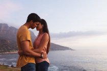 Vista lateral de una joven pareja de raza mixta de pie en una playa abrazando y a punto de besarse al atardecer - foto de stock