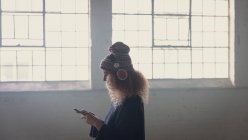 Side view of a young Caucasian woman with curly hair wearing a black long sleeves and headphones over a beanie while using a mobile phone inside an empty warehouse — Stock Photo