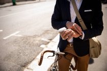 Vista frontal sección media de un hombre sosteniendo un café para llevar y comprobando la hora, sentado en su bicicleta en una calle de la ciudad. Nómada digital en movimiento . - foto de stock