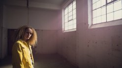 Vista laterale di una giovane donna caucasica con i capelli ricci che indossa una giacca gialla sopra una camicia grigia guardando attentamente la telecamera all'interno di un magazzino vuoto — Foto stock