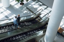 Vista de ángulo alto de hombres de negocios que interactúan entre sí mientras se mueven abajo en escaleras mecánicas en la oficina
. - foto de stock