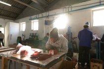 Frontansicht eines jungen afrikanisch-amerikanischen Mannes mit Hut, der an einer Werkbank sitzt und mit ausgeschnittenen roten Lederformen in einer Werkstatt einer Cricketfabrik arbeitet, im Hintergrund arbeiten Kollegen an anderen Teilen der Produktionslinie. — Stockfoto