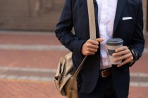 Вид спереди: стоящий на городской улице мужчина в куртке, с сумкой на плече, держащий в руках кофе. Цифровая реклама на ходу . — стоковое фото