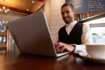 Vue de face gros plan d'un jeune homme caucasien utilisant un ordinateur portable assis à une table à l'intérieur d'un café. Nomade numérique en mouvement . — Photo de stock