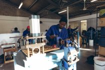 Frontansicht eines jungen afrikanisch-amerikanischen Mannes mit Hut und Arbeitskleidung, der in einer Werkstatt einer Cricketfabrik eine Maschine bedient, im Hintergrund arbeiten Kollegen an anderen Teilen der Produktionslinie. — Stockfoto
