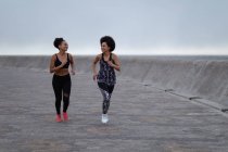 Вид спереду на двох молодих сестер змішаної раси в спортивному одязі, що бігають і дивляться один на одного, посміхаючись у зовнішньому міському просторі — стокове фото