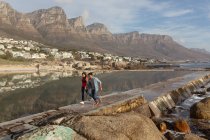 Vista frontale di una giovane coppia mista che cammina e corre su rocce in riva al mare, con cielo azzurro e montagne sullo sfondo — Foto stock