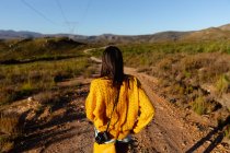 Vue arrière rapprochée d'une jeune femme métisse marchant le long d'un sentier à travers un paysage rural ensoleillé vers les montagnes à l'horizon . — Photo de stock