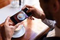Au-dessus de l'épaule de l'homme prenant des photos de son café avec un smartphone, assis à une table à l'intérieur d'un café. Nomade numérique en mouvement . — Photo de stock