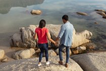Vista trasera elevada de una joven pareja de mestizos tomados de la mano sobre rocas junto al mar - foto de stock