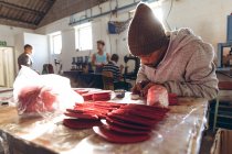 Vue de face gros plan d'un jeune Afro-Américain portant un chapeau assis sur un établi travaillant avec des formes découpées de cuir rouge dans un atelier d'une usine fabriquant des balles de cricket — Photo de stock