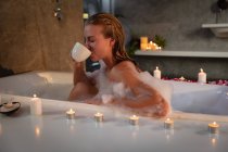Close up side view of a young Caucasian woman sitting in a foam bath with lit candles around it drinking a cup of tea. — Stock Photo