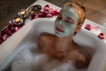 Close up front view of a young Caucasian woman lying back in a bath with a face mask, with petals and lit candles around the bathtub. — Stock Photo