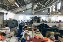 Вид сбоку на группу женщин-коллег, работающих на швейных машинах и сортирующих ткани на фабрике спортивной одежды, с другими коллегами, работающими на заднем плане . — стоковое фото