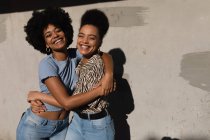 Retrato de duas jovens adultas mestiças irmãs de pé contra uma parede abraçando-se com os braços ao redor uns dos outros, olhando para a câmera e sorrindo ao sol — Fotografia de Stock