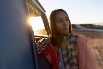 Vue de face rapprochée d'une jeune femme métissée réfléchie avec une couverture sur les épaules, penchée sur la porte d'une camionnette et regardant loin le coucher du soleil lors d'un arrêt sur une route — Photo de stock