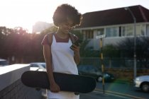 Вид спереди на молодую женщину смешанной расы, стоящую на городской улице, держащую в руках скейтборд и использующую смартфон, подсвеченный солнечным светом — стоковое фото