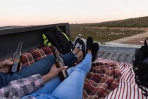 Feche a seção baixa das pernas do casal sentado na parte de trás de seu caminhão pick-up segurando garrafas de cerveja durante uma parada em uma viagem de carro . — Fotografia de Stock