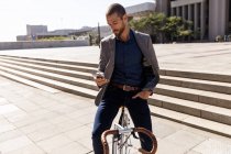 Vista frontal de perto de um jovem caucasiano usando um smartphone, sentado em sua bicicleta em uma rua da cidade. Digital Nomad em movimento . — Fotografia de Stock