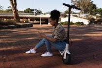 Вид сбоку улыбающейся молодой женщины смешанной расы, сидящей на электрическом скутере в городском парке с помощью смартфона — стоковое фото