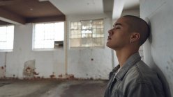 Вид сбоку на молодого латиноамериканца с пирсингом в серой куртке, прислонившегося к белой стене внутри пустого склада, смотрящего в сторону от камеры — стоковое фото