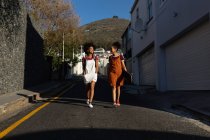 Vista frontal de dos hermanas adultas jóvenes de raza mixta, una con una mochila y la otra con un monopatín, hablando y sonriendo mientras caminan en una calle bajo el sol - foto de stock