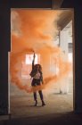 Vorderansicht einer jungen kaukasischen Frau mit lockigem Haar, die eine Lederjacke trägt, während sie aufmerksam in die Kamera blickt und eine Rauchmaschine in der Hand hält, die einen orangefarbenen Rauch in einer leeren Lagerhalle erzeugt — Stockfoto