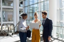 Front view of young Caucasian businesswoman standing between a young African American and a young Caucasian businessmen looking at a laptop in a modern office lobby. — Stock Photo