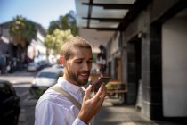 Side view close up of a smiling young Caucasian man talking on a smartphone holding it in front of his face, standing in a city street. Digital Nomad on the go. — Stock Photo