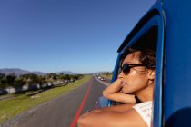 Nahaufnahme einer jungen Frau mit Sonnenbrille, die sich aus der Beifahrerseite eines Pick-ups lehnt, während dieser auf einer Autoreise die Autobahn hinunterfährt — Stockfoto