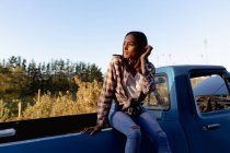 Vista frontal de una joven mujer de raza mixta sentada en la parte trasera de una camioneta mirando hacia otro lado, durante una parada en un viaje por carretera . - foto de stock