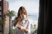 Vista frontal de cerca de una joven mujer caucásica con una camisa blanca de pie en un balcón bebiendo una taza de café y mirando hacia abajo, palmeras y playa en el fondo . - foto de stock