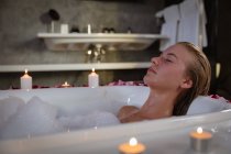 Закройте боковой вид на молодую кавказку, расслабляющуюся в пенной ванне с зажженными свечами вокруг нее с закрытыми глазами . — стоковое фото