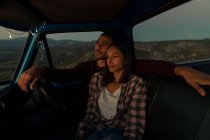 Vista lateral de cerca de una pareja de jóvenes y reflexivos de raza mixta sentados en su camioneta, descansando y mirando hacia la vista al atardecer, durante una parada en un viaje por carretera . - foto de stock