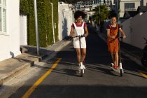Frontansicht von zwei jungen erwachsenen Mischlingsschwestern, die lächelnd Roller in einer Straße in der Sonne fahren — Stockfoto