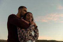 Side view of a young mixed race couple sitting outside embracing at dusk during a stop off on a road trip. The woman is smiling and has her eyes closed. — Stock Photo