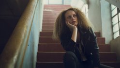 Низький кут зору молода кавказька жінка з Кучеряве волосся в шкіряній куртці сидить на сходах з рук на підборіддя — стокове фото
