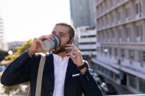 Vue de face gros plan d'un jeune homme caucasien buvant un café à emporter et parlant sur son smartphone, debout dans la ville. Nomade numérique en mouvement . — Photo de stock